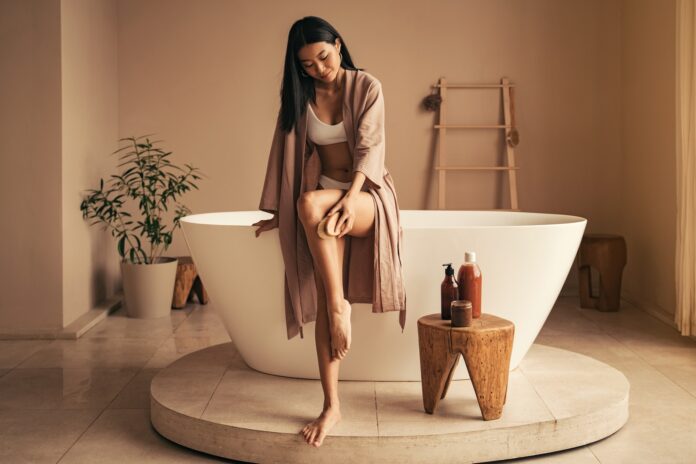 Young asian woman doing peeling procedures, dry brushing with wooden brush in the bathroom